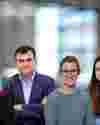 A group of six adults standing side by side in a bright indoor setting. They are smiling and dressed in casual and smart-casual clothing, with a blurred office background behind them.