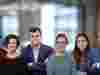 A group of six adults standing side by side in a bright indoor setting. They are smiling and dressed in casual and smart-casual clothing, with a blurred office background behind them.